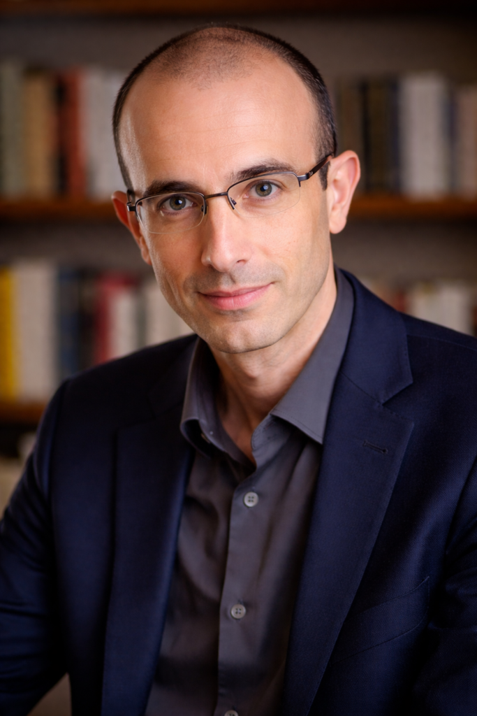 Realistic portrait of Yuval Noah Harari, author of Sapiens, wearing a navy blazer and light blue shirt, standing in front of a blurred bookshelf, professional and thoughtful expression.